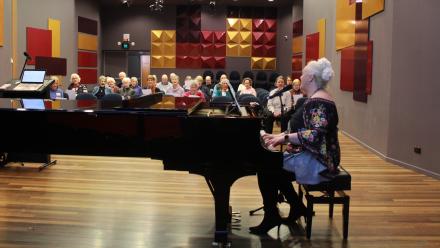 A person playing piano with an audience of senior adults singing and seated in the background